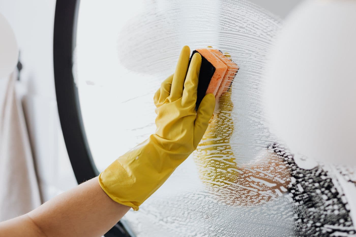 Gloved hand cleaning a mirror with sponge and soapy foam
